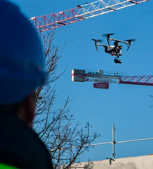 Drone professionnel en vol sur un chantier de construction, surveillé par un opérateur en casque de sécurité avec grues en arrière-plan.
