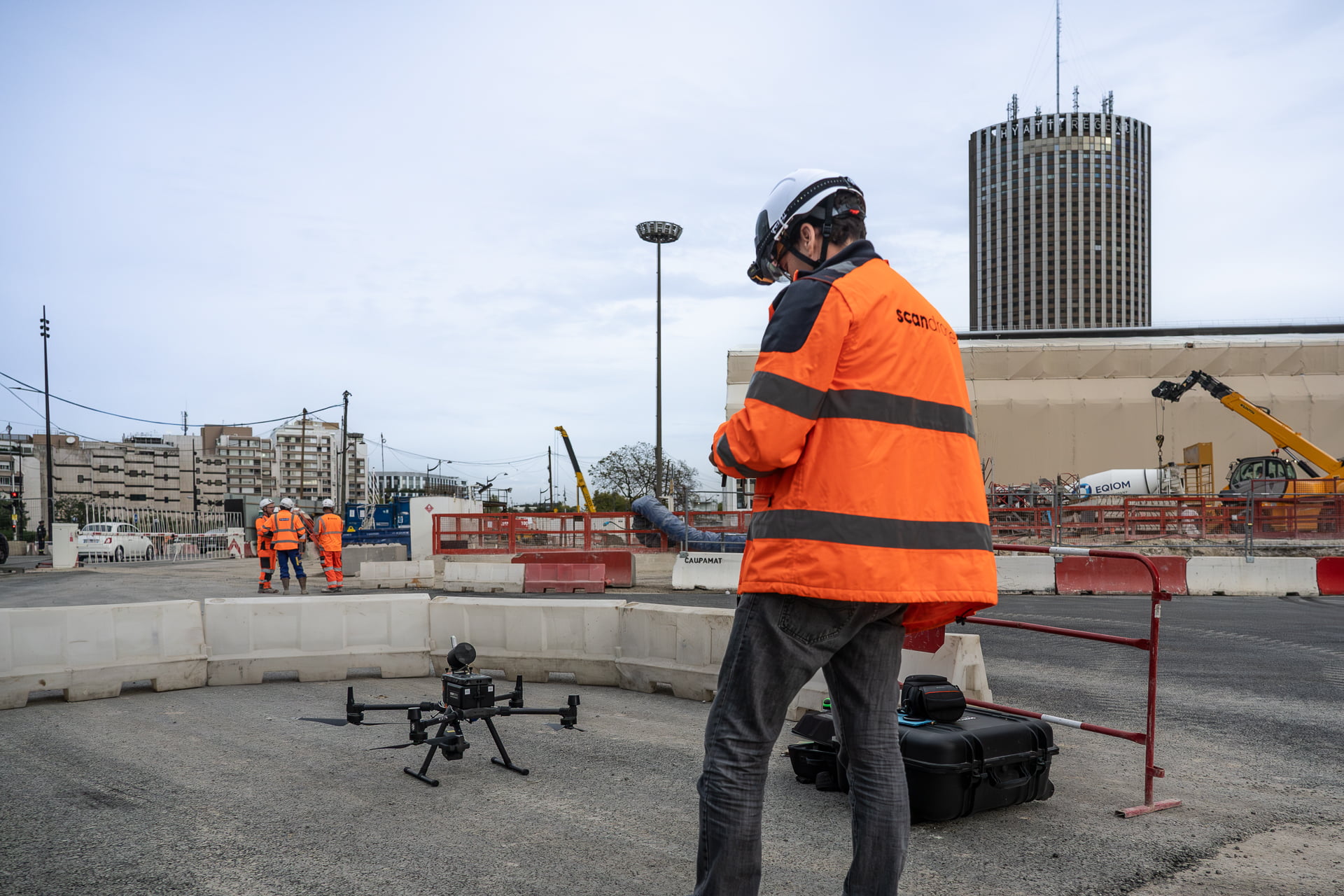 Opérateur Scandrone en veste orange et casque, pilotant un drone Matrice 300 RTK équipé de capteurs sur une zone de chantier urbain, avec en arrière-plan des engins de construction et une équipe en intervention. Entreprise de drones professionnels sur un chantier.