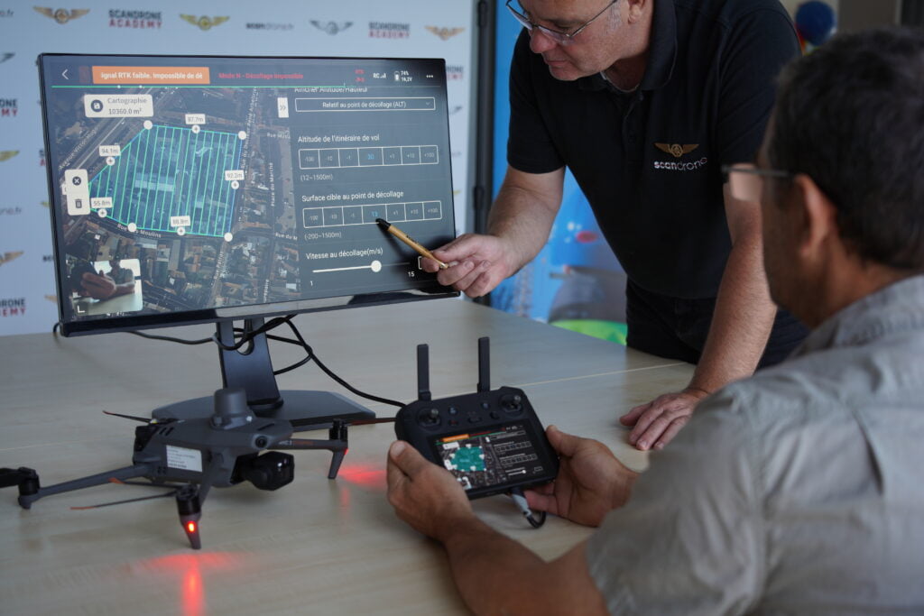 Deux opérateurs Scandrone en session de planification de mission photogrammétrique, avec un drone DJI Mavic 3 Enterprise posé sur la table, un moniteur affichant le plan de vol et une radiocommande en main.