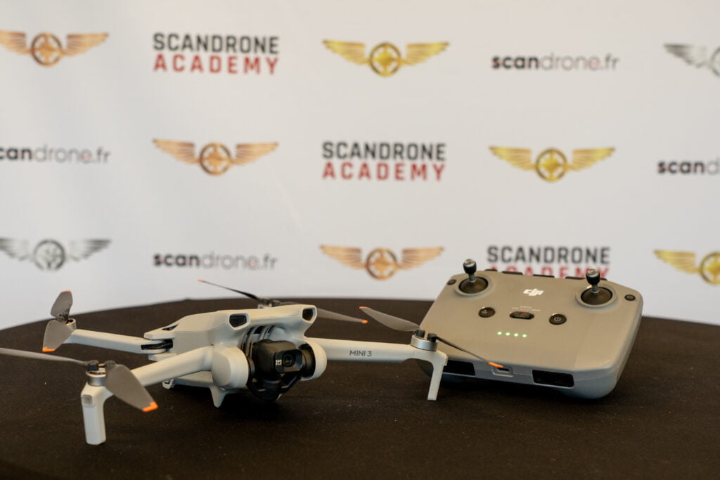 Drone DJI Mini 3 posé sur une table aux côtés de sa télécommande, devant un fond aux logos de Scandrone Academy et scandrone.fr