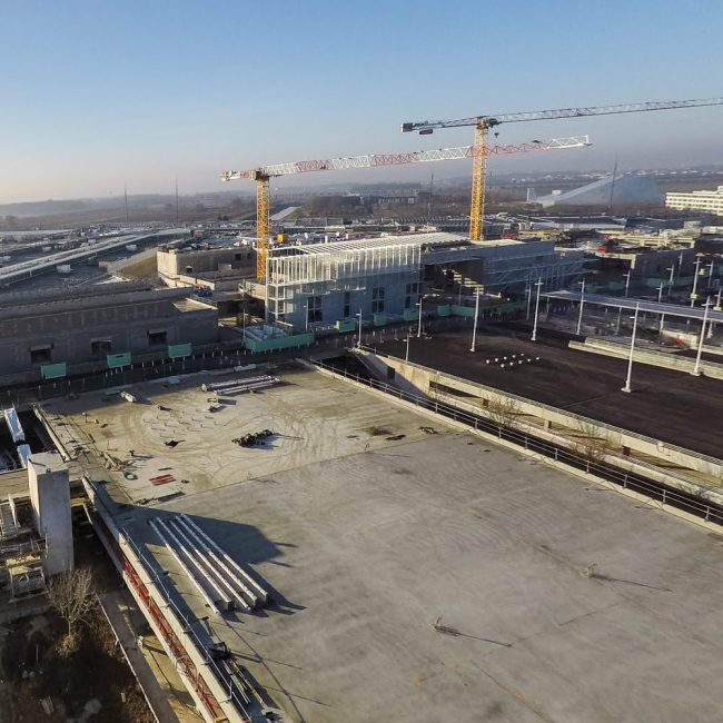 Vue aérienne chantier Vue aérienne d’un chantier de grande envergure avec grues, bâtiment en construction et dalle en béton, illustrant le suivi de projet et le contrôle d’avancement par drone dans un contexte de travaux publics.